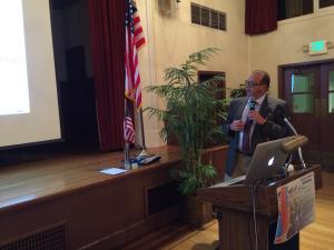 A speaker holds a microphone beside a lectern, laptop, and projection screen in the parish hall.