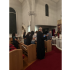 Participants stand beside wooden pews in St. John's sanctuary, with a black-robed clergyman in the foreground.