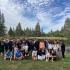 A group of young people and adults poses on the grass beside a lake surrounded by pine trees at Hye Camp.
