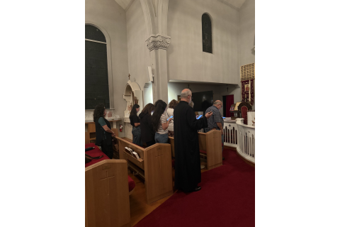 Participants stand beside wooden pews in St. John's sanctuary, with a black-robed clergyman in the foreground.