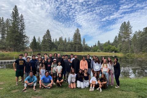 A group of young people and adults poses on the grass beside a lake surrounded by pine trees at Hye Camp.