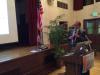 A speaker holds a microphone beside a lectern, laptop, and projection screen in the parish hall.